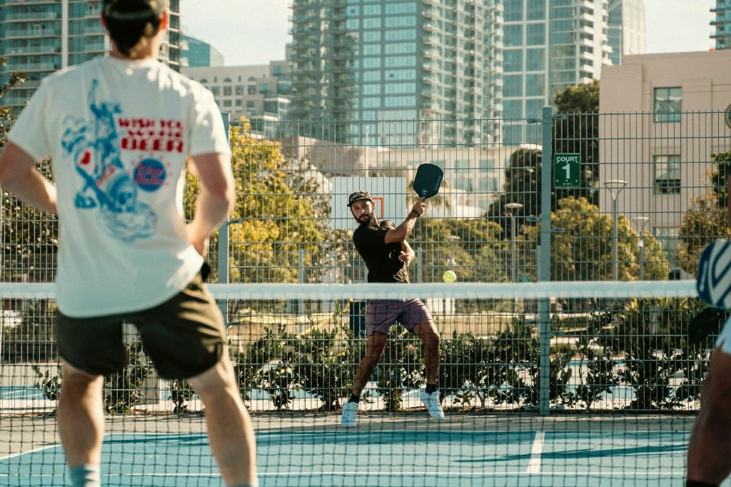 Pickleball player swinging a paddle on an outdoor blue court with a city skyline behind the fence