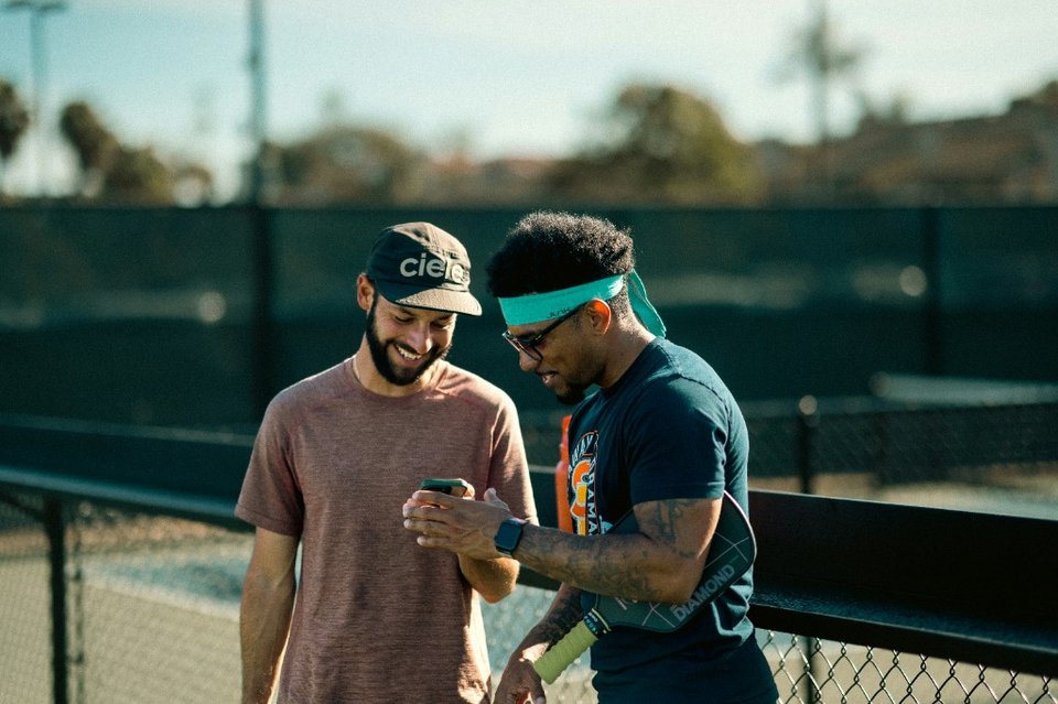 Two tennis players reviewing a phone together beside an outdoor court fence